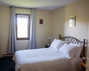 The sunlight shines through the window of a St. Charles Retreat Centre bedroom featuring a comfortable bed with pillows comforter and a nightstand with lamp.