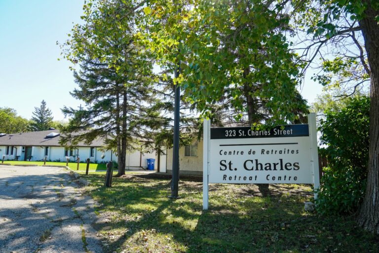 The large entrance sign of the St. Charles Retreat Centre with sunlight flowing through the trees and the centre entrance is in the background.